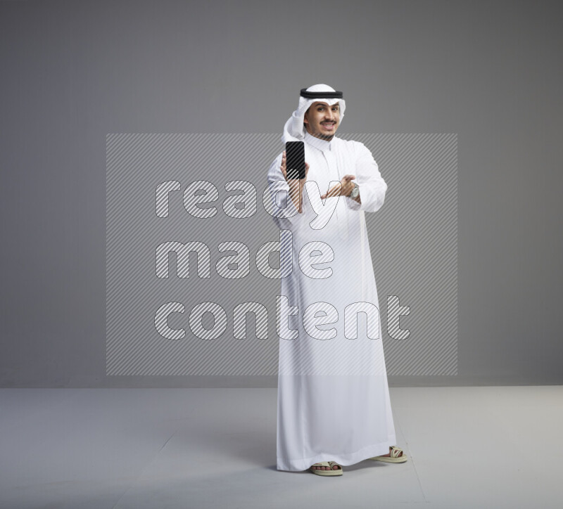 A Saudi man standing wearing thob and white shomag showing phone to camera on gray background