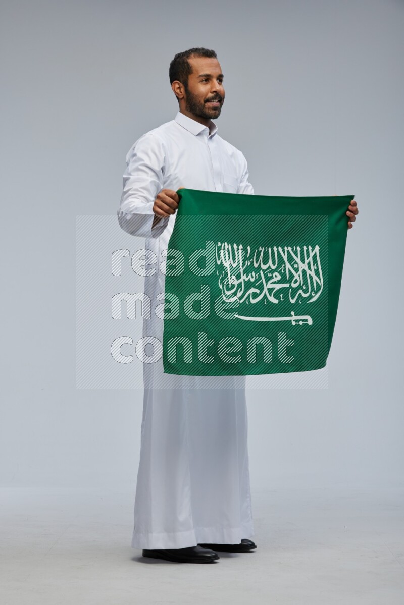 Saudi man wearing Thob standing holding Saudi flag on Gray background