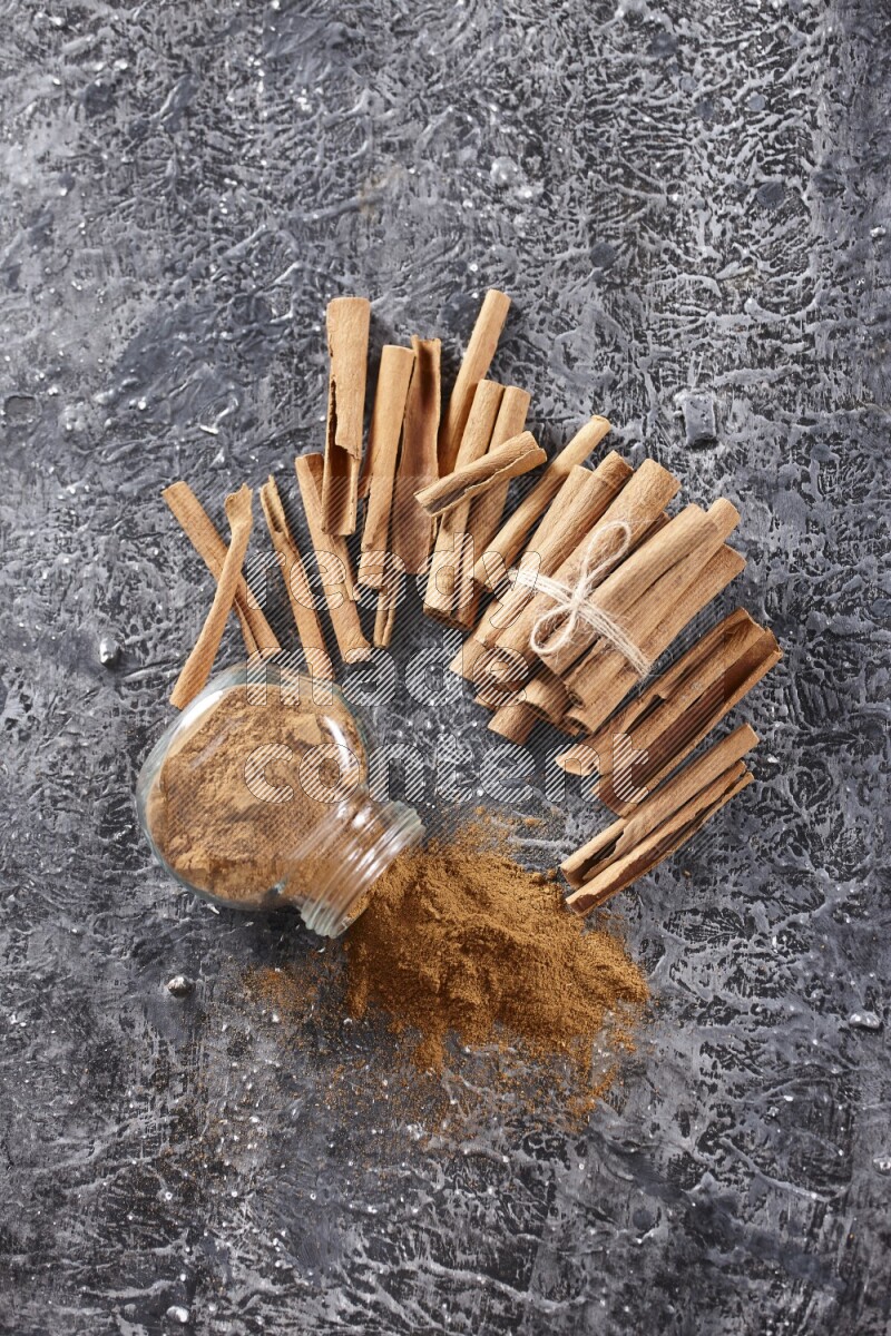 Herbal glass jar full of cinnamon powder flipped with cinnamon sticks stacked and bounded on a textured black background