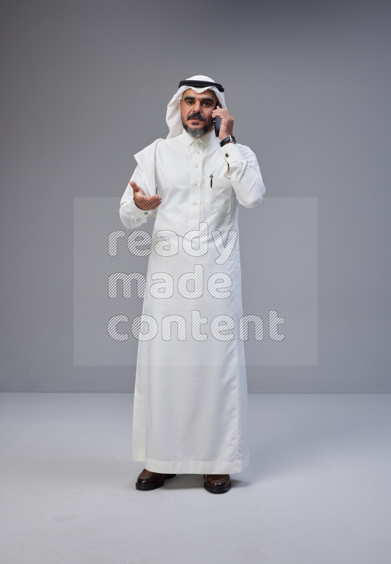 Saudi man Wearing Thob and white Shomag standing talking on phone on Gray background