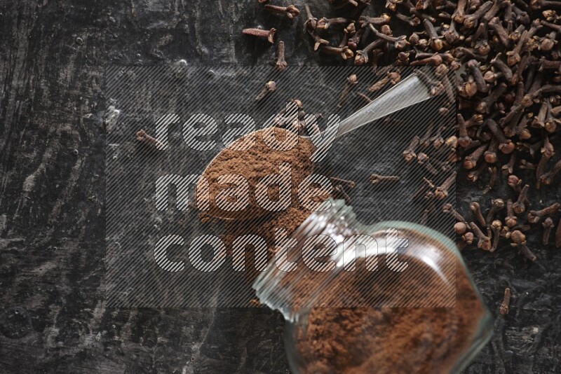 A flipped glass spice jar and a metal spoon full of cloves powder with cloves spread on textured black flooring