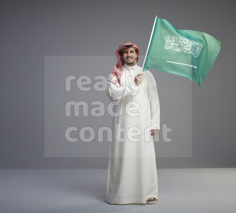A Saudi man standing wearing thob and red shomag raising big Saudi flag on gray background