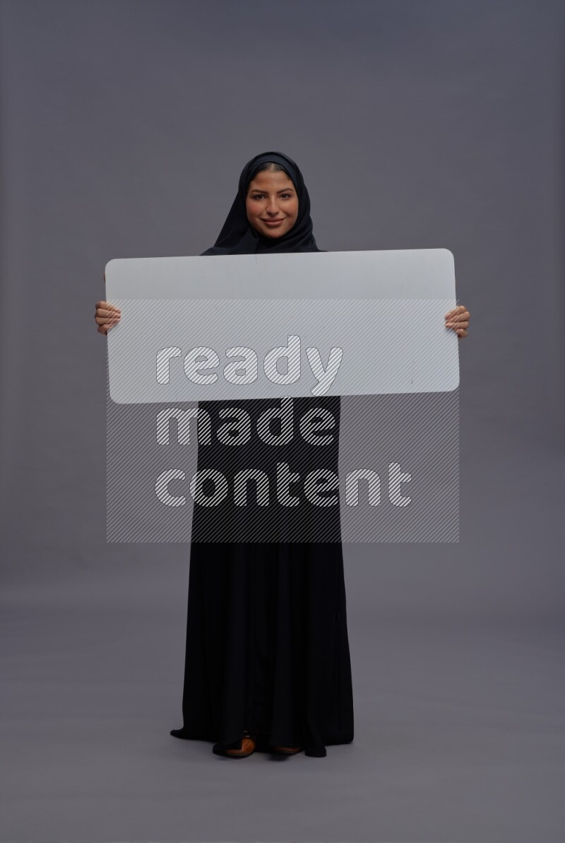 Saudi woman wearing Abaya standing holding white board on gray background