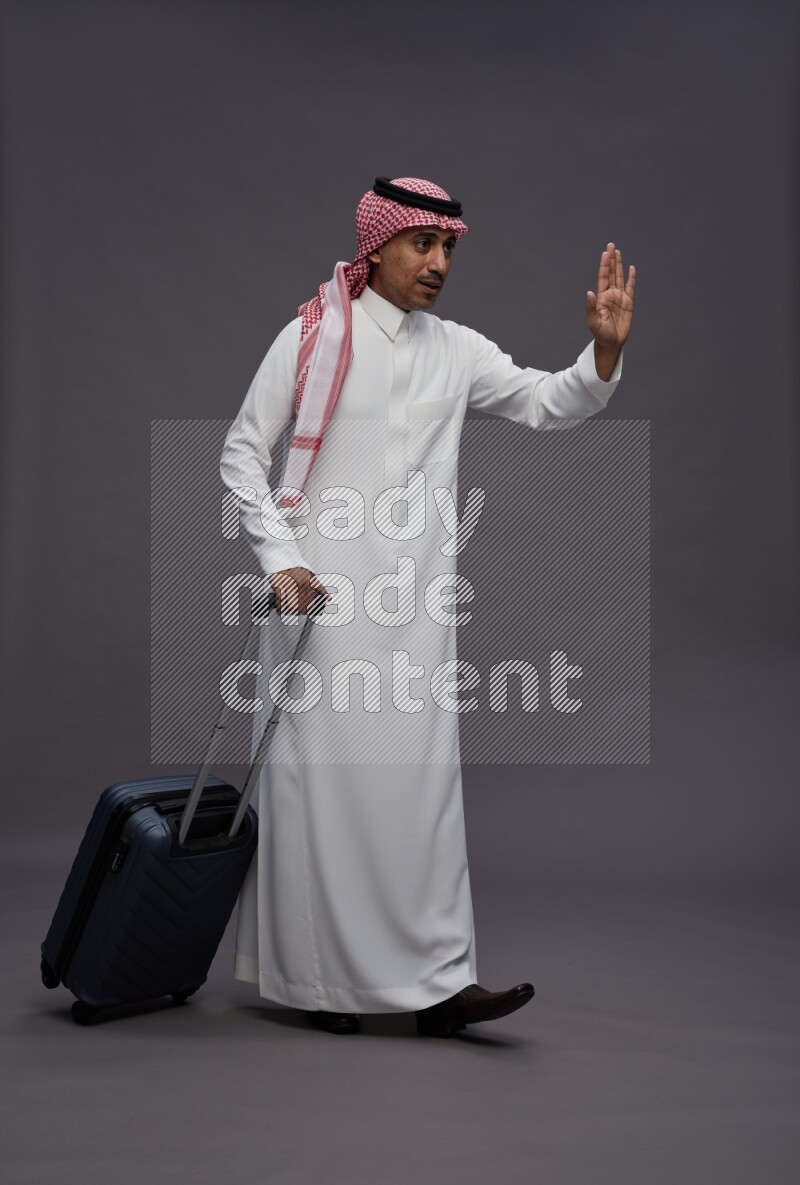 Saudi man wearing thob and shomag standing holding bag on gray background