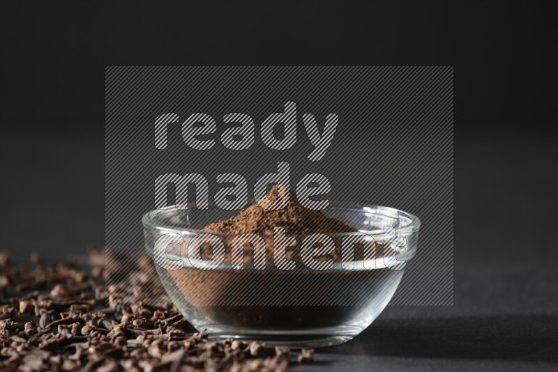 A glass bowl full of cloves powder with cloves grains spread on black flooring