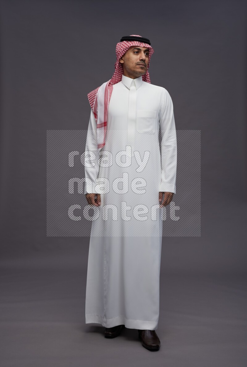 Saudi man wearing thob and shomag standing interacting with the camera on gray background