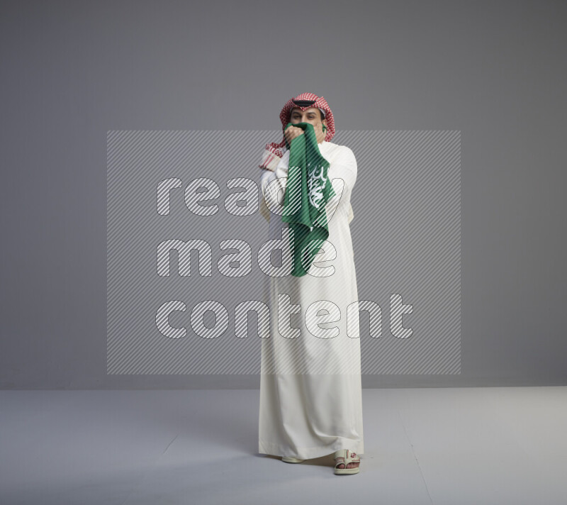 A Saudi man standing wearing thob and red shomag kissing big Saudi flag on gray background