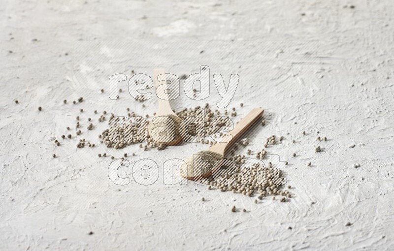 2 wooden spoons full of white pepper powder with white pepper beads on textured white flooring