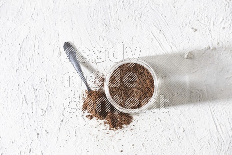 A glass jar full of cloves powder with a metal spoon on a textured white flooring