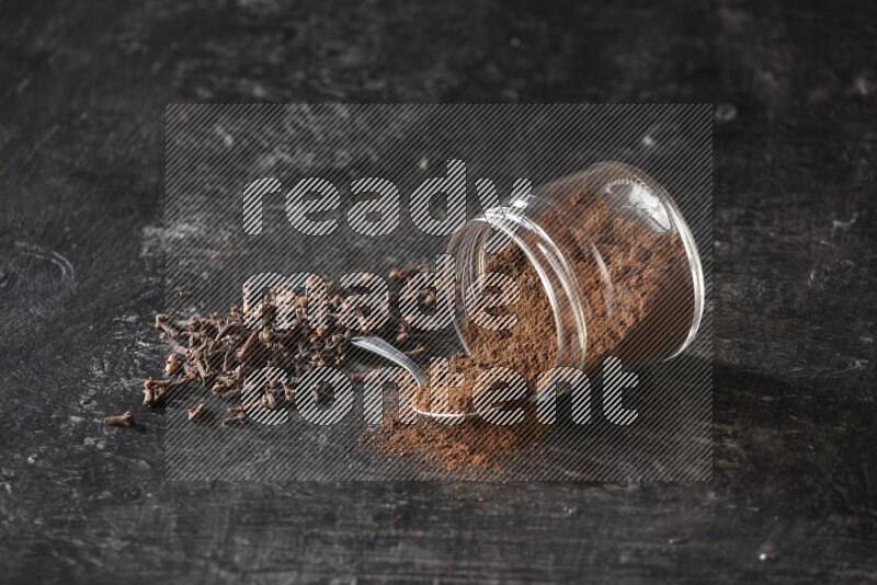 A flipped glass jar and metal spoon full of cloves powder with cloves spread on a textured black flooring