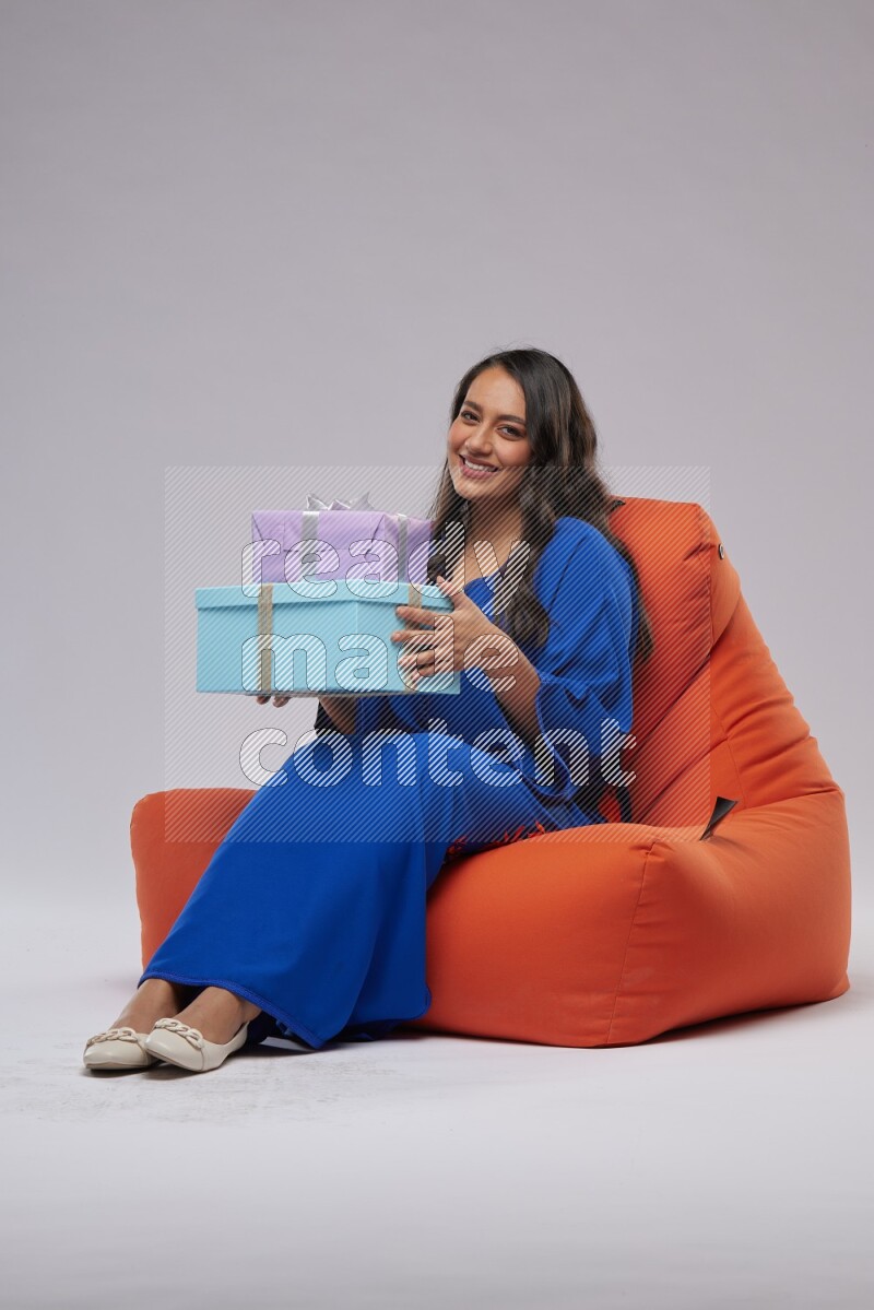 A woman sitting on an orange beanbag wearing Jalabeya holding a gift box