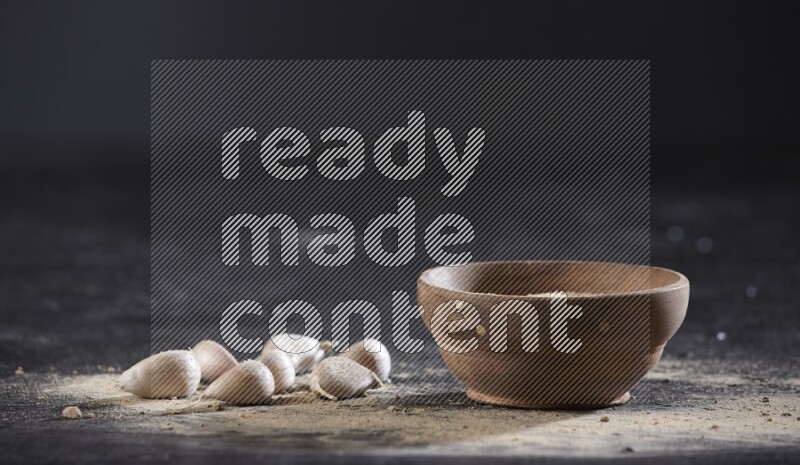 A wooden bowl full of garlic powder with some garlic cloves beside it on a textured black flooring