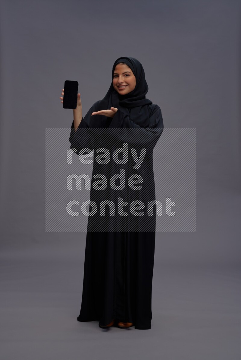Saudi woman wearing Abaya standing showing phone to camera on gray background