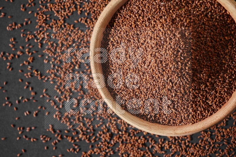 A wooden bowl full of garden cress seeds surrounded by seeds on a black flooring