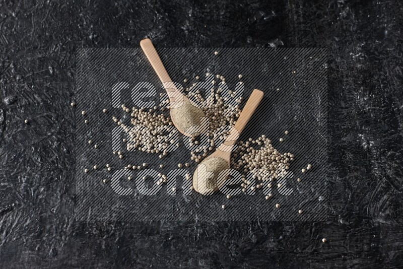 2 wooden spoons full of white pepper powder with white pepper beads on textured black flooring