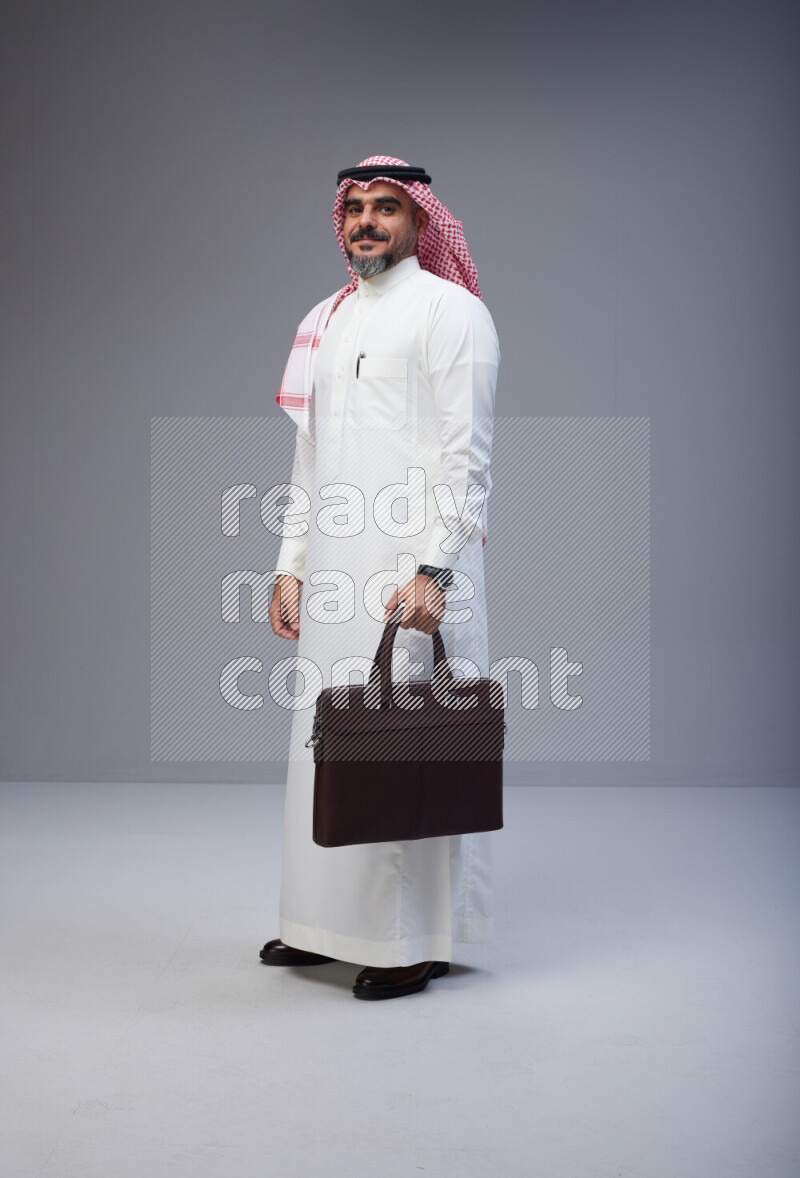 Saudi man Wearing Thob and red Shomag standing holding bag on Gray background