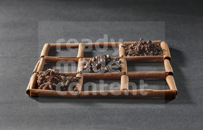 9 squares of cinnamon sticks full of coffee beans in the middle surrounded by nutmeg, dried mint, cloves, dried basil, dried ginger, cinnamon, star anise and cardamom on black flooring