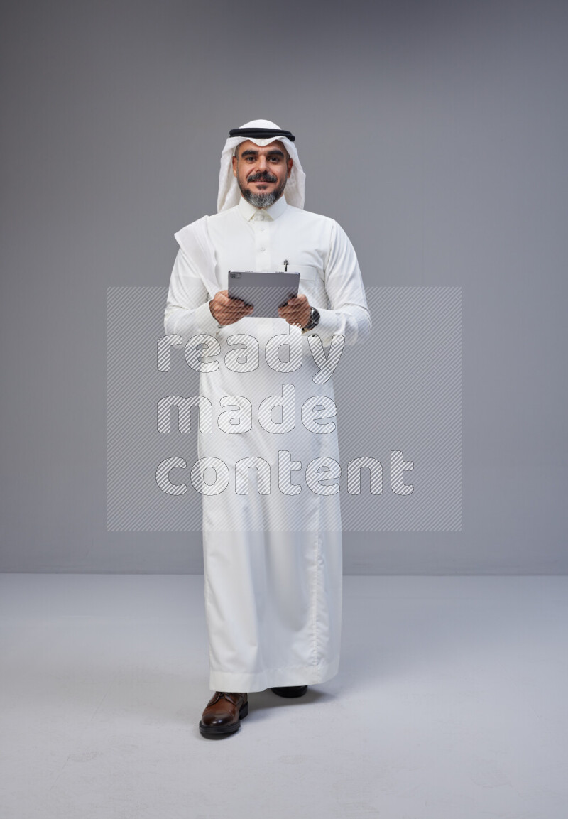 Saudi man Wearing Thob and white Shomag standing working on tablet on Gray background