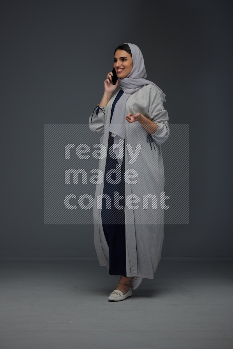 A Saudi woman wearing a light gray Abaya and head scarf standing and talking in the phone eye level on a grey background