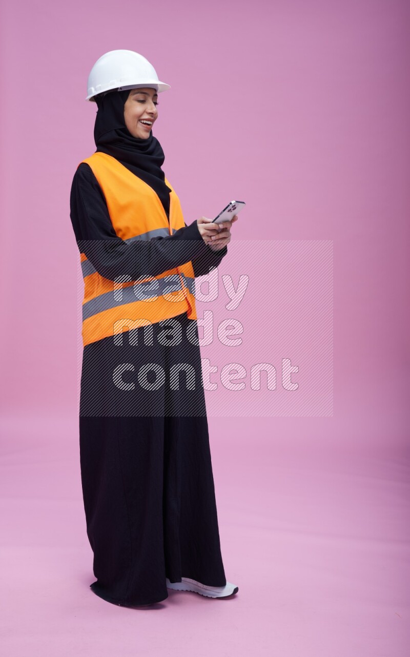 Saudi woman wearing Abaya with engineer vest and helmet standing texting on phone on pink background