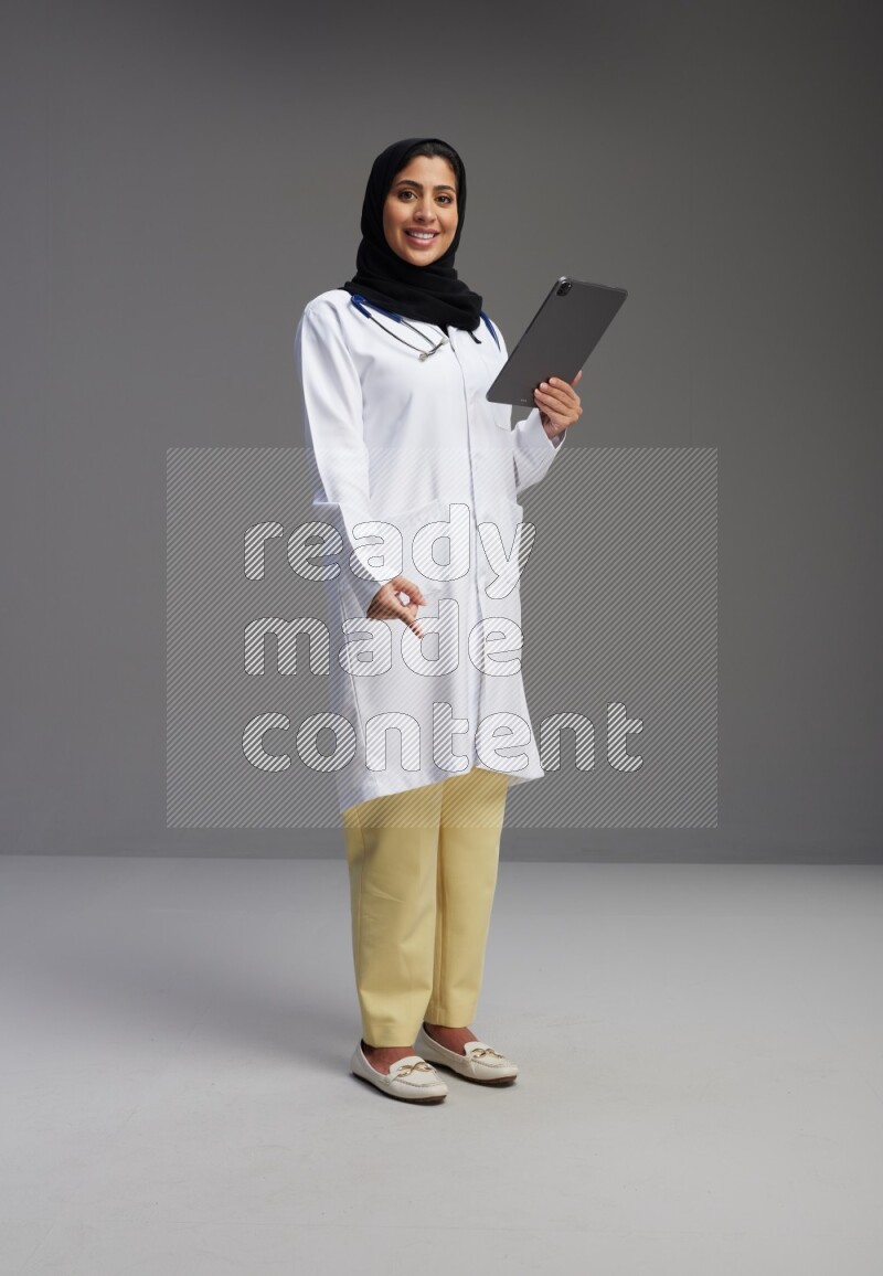 Saudi woman wearing lab coat with stethoscope standing working on tablet on Gray background