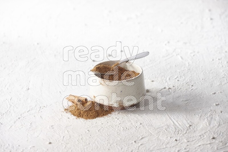 Ceramic beige bowl full of cinnamon powder and a metal spoon with cinnamon sticks next of it on a textured white background