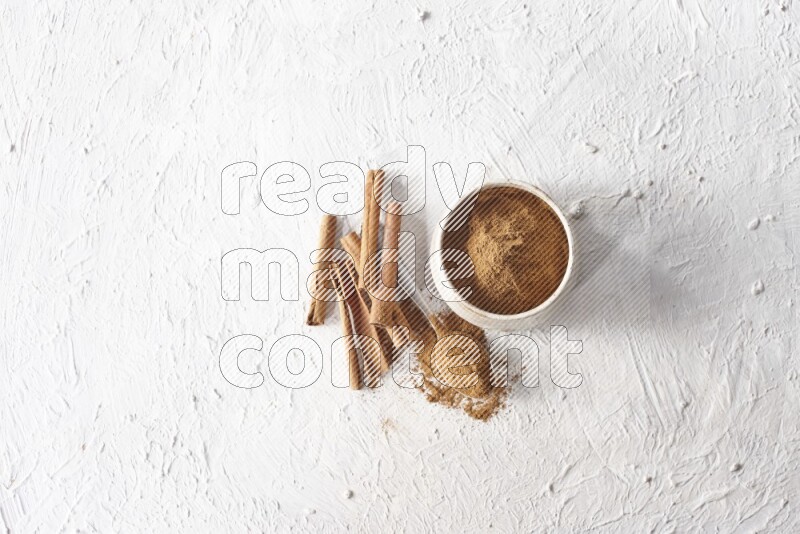Ceramic beige bowl full of cinnamon powder and a metal spoon with cinnamon sticks next of it on a textured white background