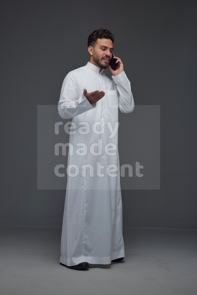 A Saudi man wearing Thobe and talking in the phone while standing and making different poses eye level on a gray background