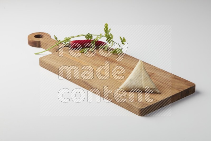 One uncooked samosa with a single red pepper and parsley on a wooden cutter on a white background