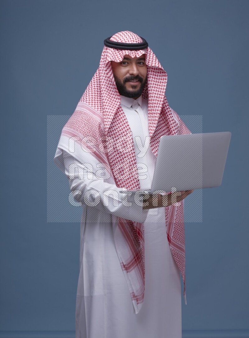 A man standing  with a laptop on Blue Background wearing Saudi Thob and Shomag