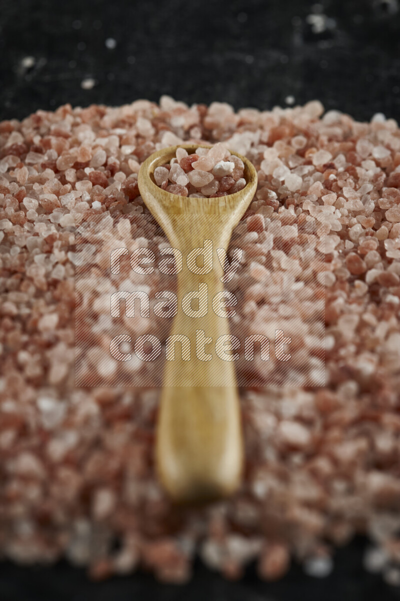A wooden spoon full of coarse himalayan salt crystals on a bunch of the crystals on black background