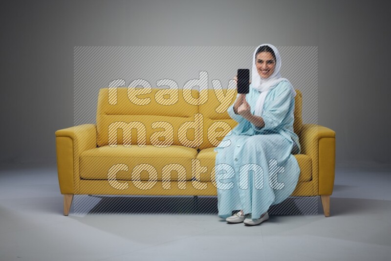 A Saudi woman wearing a light blue Abaya and white head scarf sitting on a yellow sofa and showing her phone's screen vertically and horizontally eye level on a grey background