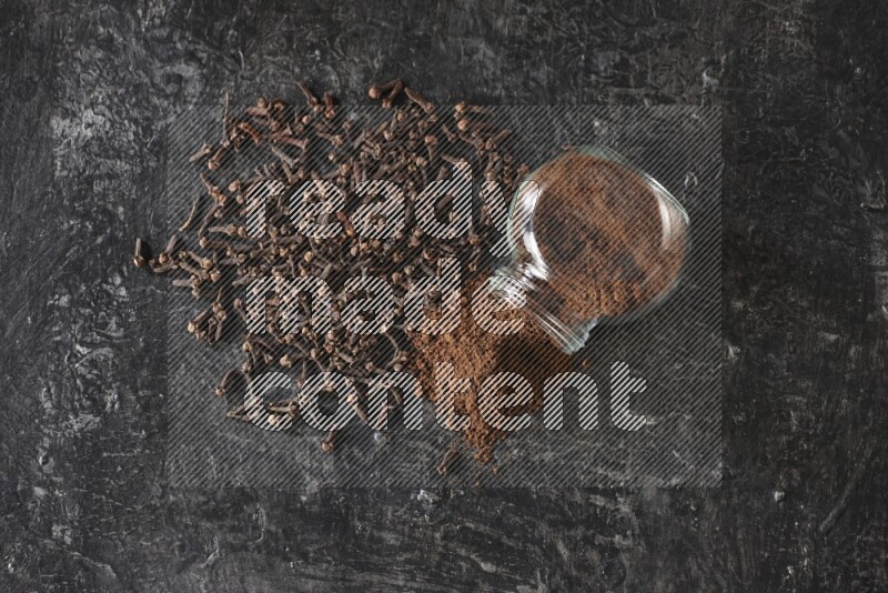 A flipped glass spice jar full of cloves powder and powder came out of it with cloves spread on textured black flooring