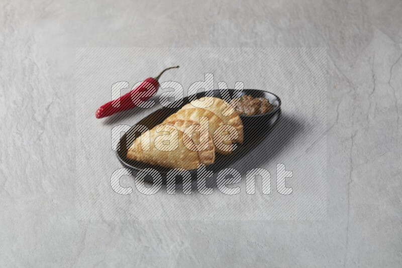 Three fried sambosas in an oval shaped black plate and a sauce in a black round ramekin on a gray background