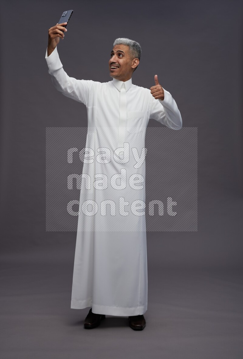 Saudi man wearing thob standing taking selfie on gray background