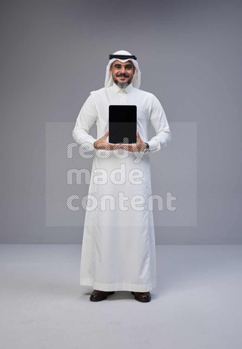 Saudi man Wearing Thob and white Shomag standing showing tablet to camera on Gray background