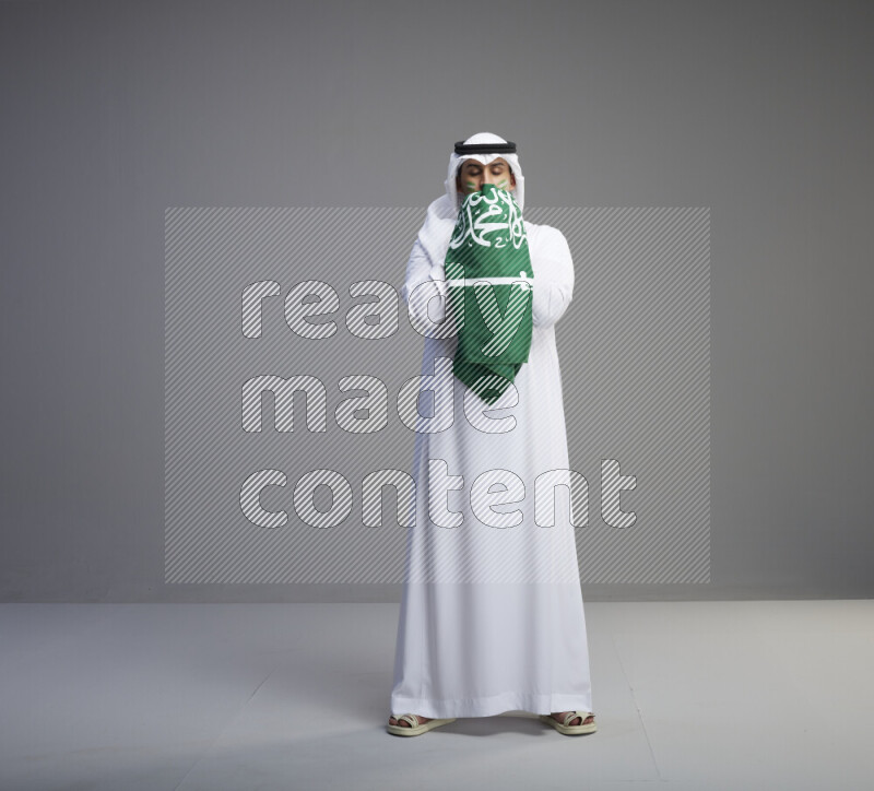A Saudi man standing wearing thob and white shomag with face painting kissing big Saudi flag on gray background