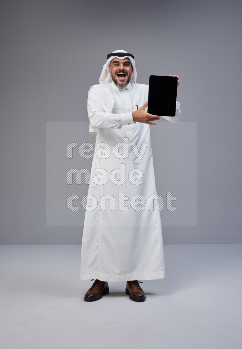 Saudi man Wearing Thob and white Shomag standing showing tablet to camera on Gray background