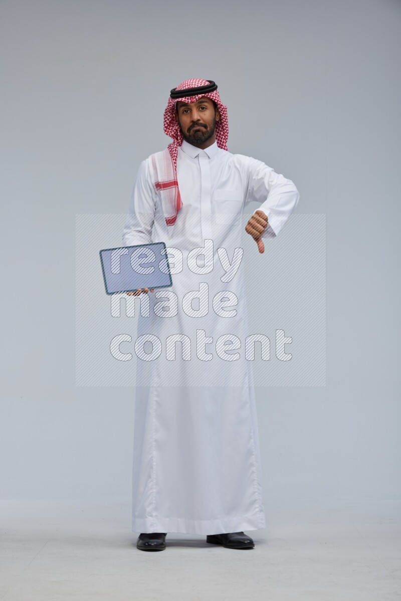 Saudi man Wearing Thob and shomag standing showing tablet to camera on Gray background