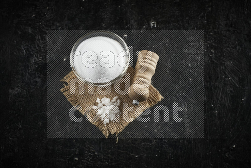 A glass bowl full of white salt with a wooden grinder on a burlap fabric all on black background