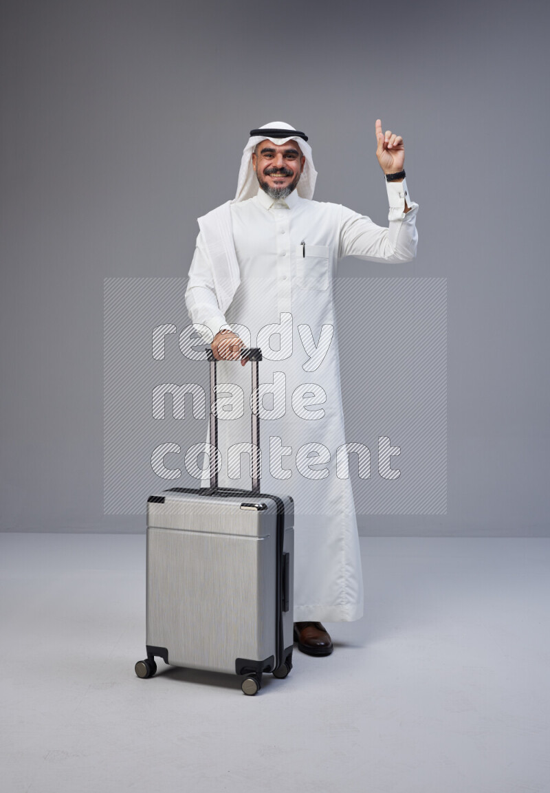 Saudi man wearing Thob and white Shomag standing holding Travel bag on Gray background