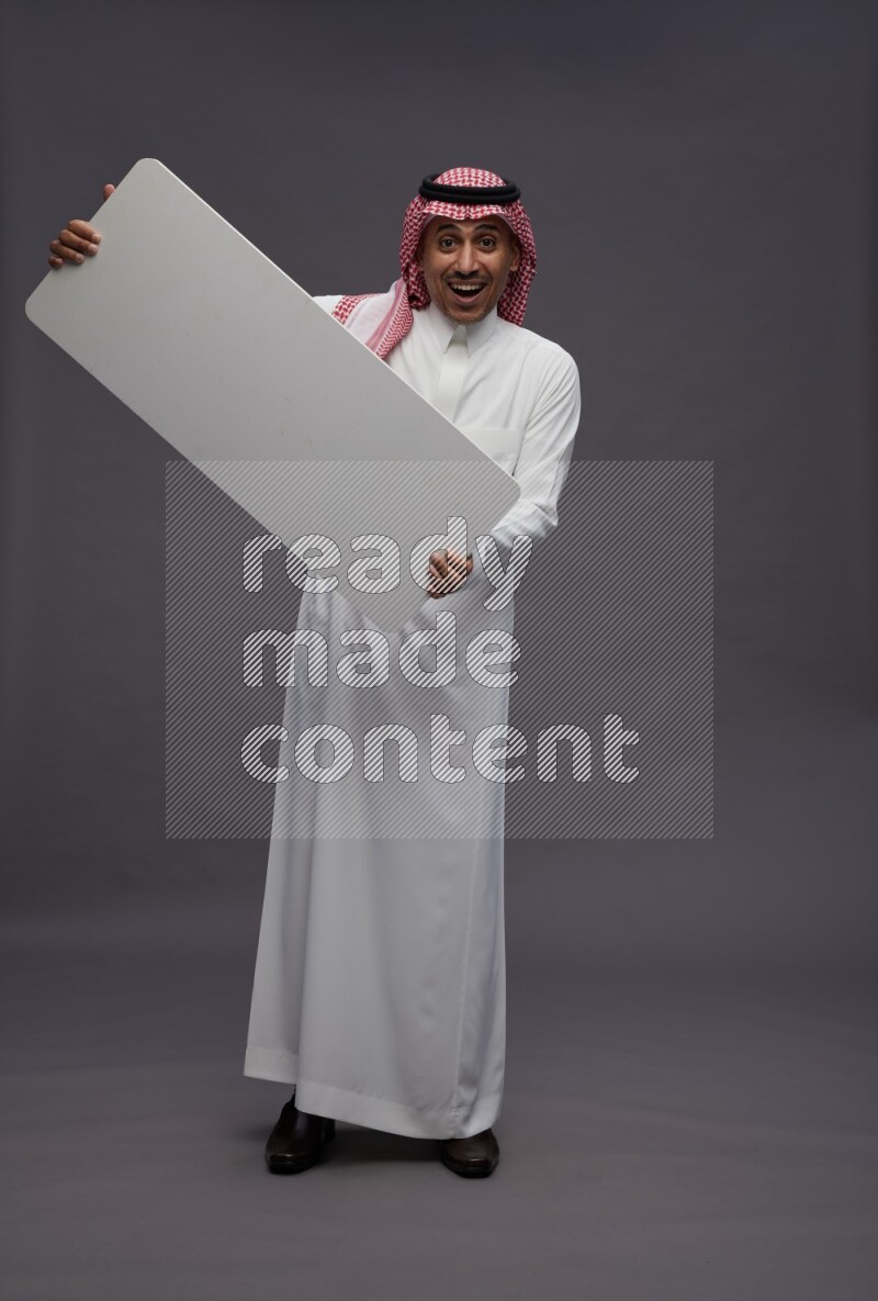 Saudi man wearing thob and shomag standing holding board on gray background
