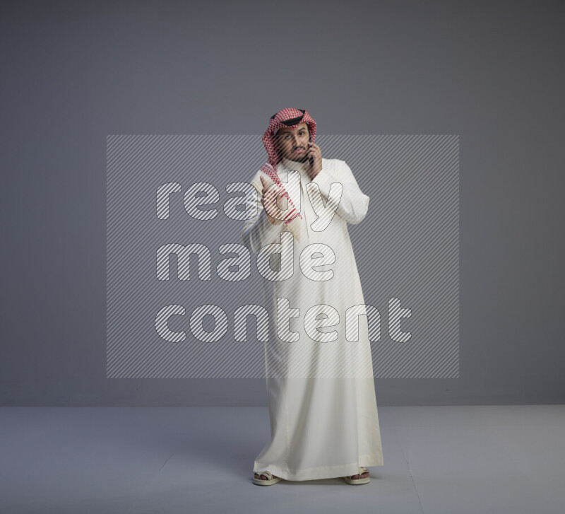 A Saudi man standing wearing thob and red shomag talking on phone on gray background