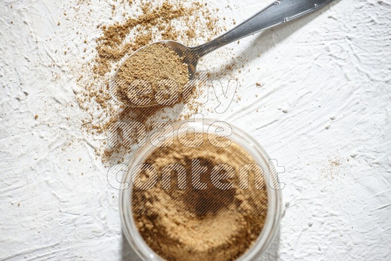 A glass jar and a metal spoon full of cumin powder on textured white flooring
