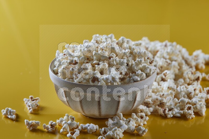 A white pottery bowl full of popcorn with popcorn beside it on a yellow background in different angles