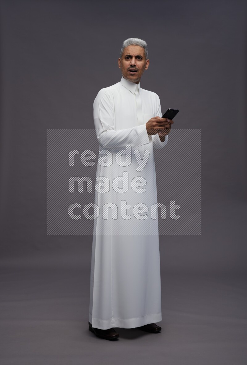 Saudi man wearing thob standing texting on phone on gray background