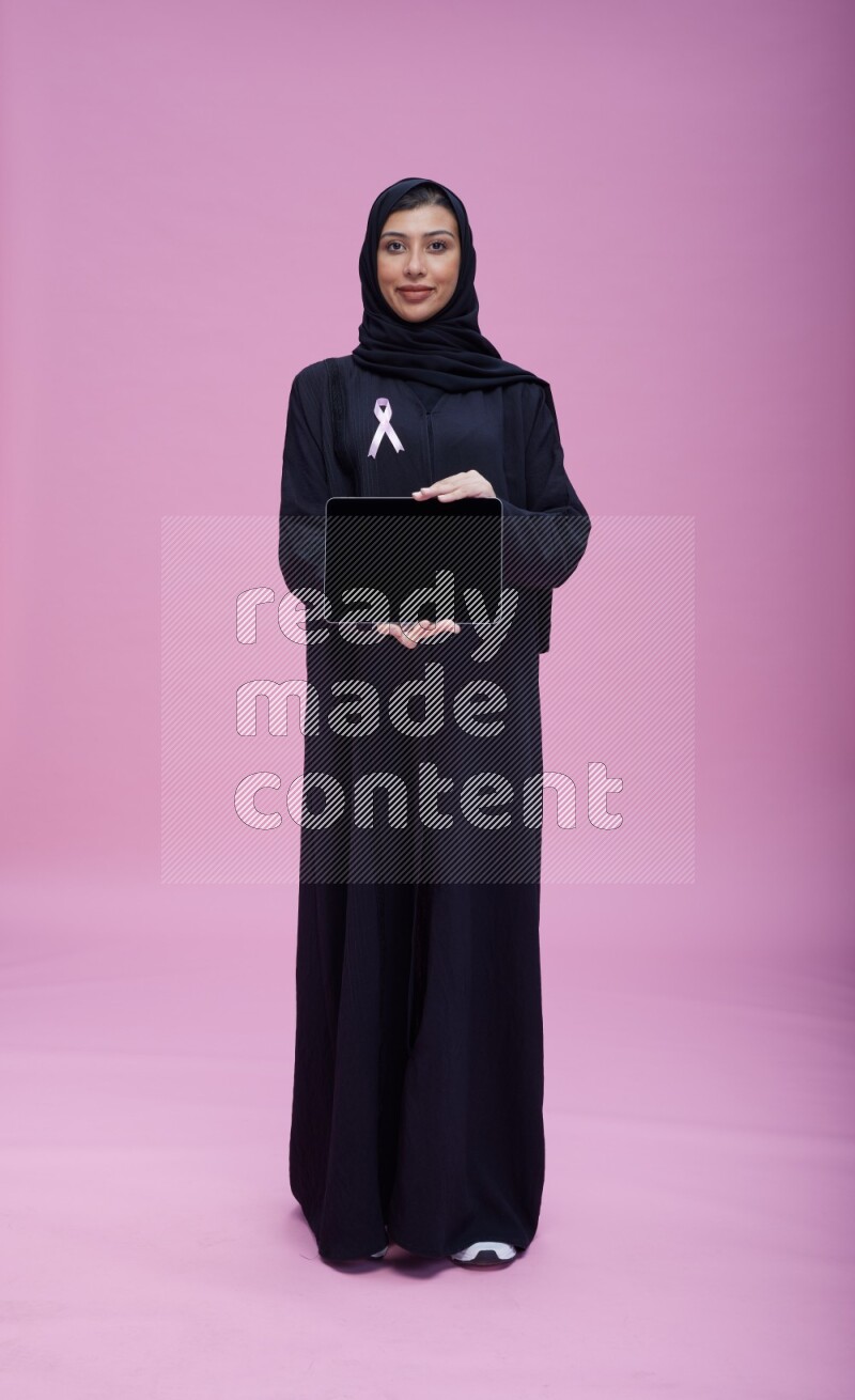 Saudi woman wearing pink ribbon on Abaya standing showing a tablet to camera on pink background