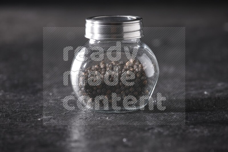 A glass spice jar full of black pepper beads on textured black flooring