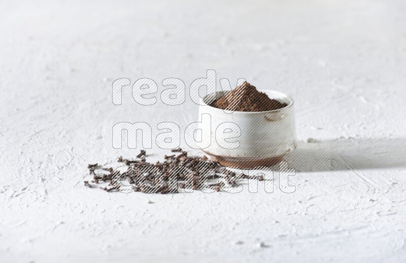 A beige ceramic bowl full of cloves powder and whole cloves on a white flooring