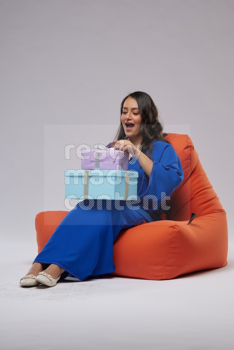 A woman sitting on an orange beanbag wearing Jalabeya holding a gift box